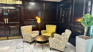 Eine Lobby mit dunklen Holzpaneelen, einem Schild mit der Aufschrift "John's Suite" und einer Sitzecke mit gemusterten Sesseln und einem runden Tisch.
