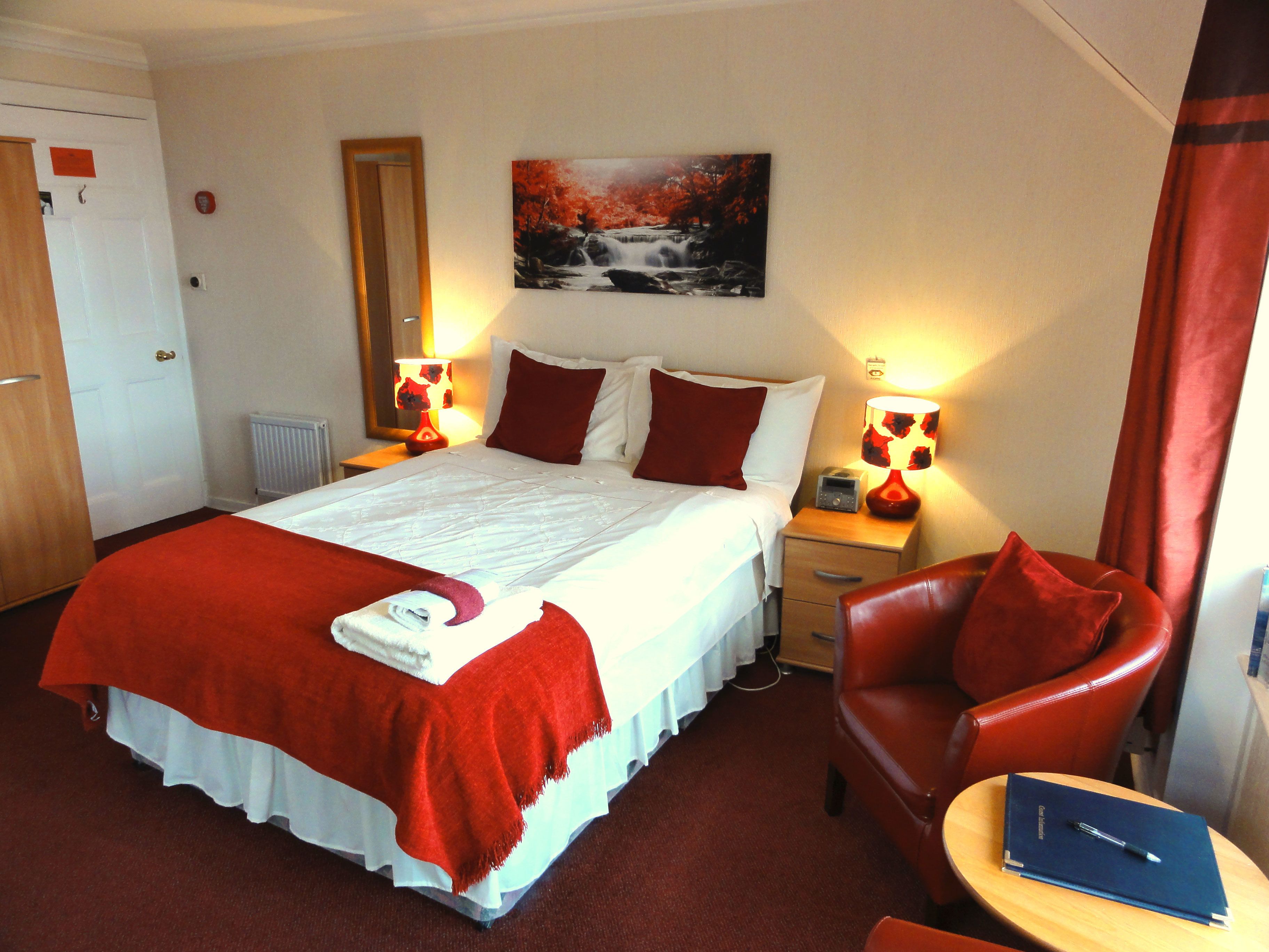 A hotel room with a large bed, red and white bedding, two bedside lamps, a red chair, a small table with a book and pen, and a painting on the wall.