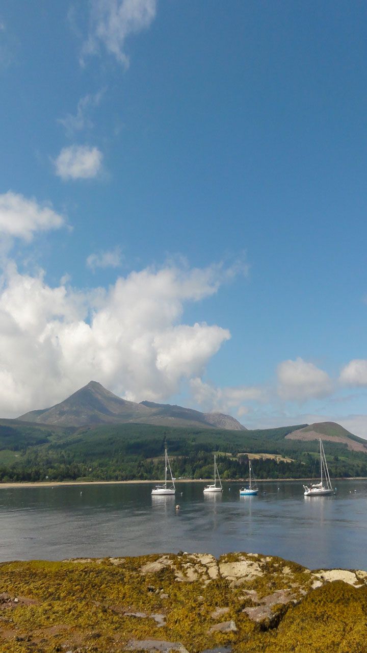 A calm lake with sailboats, a mountain in the background, and a rocky shoreline.