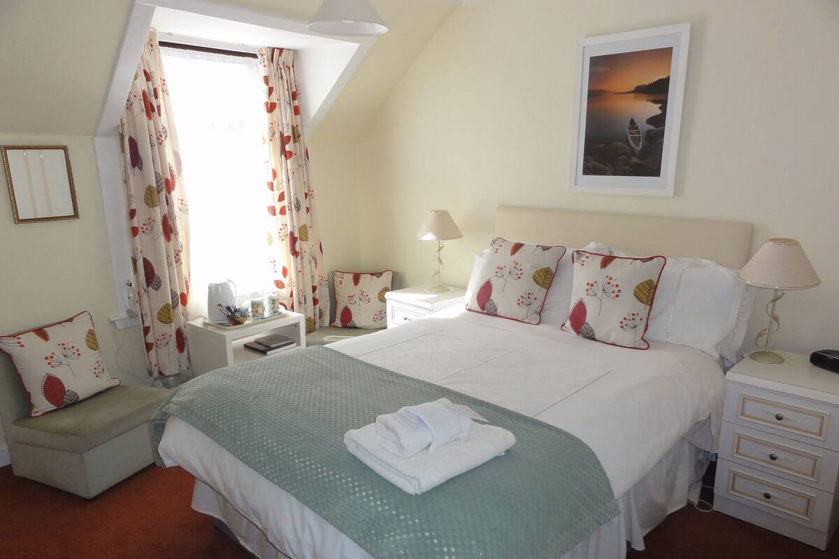 A neatly arranged bedroom with a bed, bedside tables, a nightstand, a window with curtains, and a framed picture on the wall.
