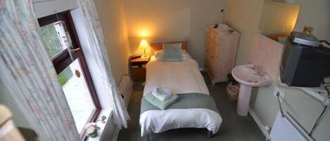 A small, cozy bedroom with two single beds, a bedside table with a lamp, a dresser, a mirror, and a window with curtains.