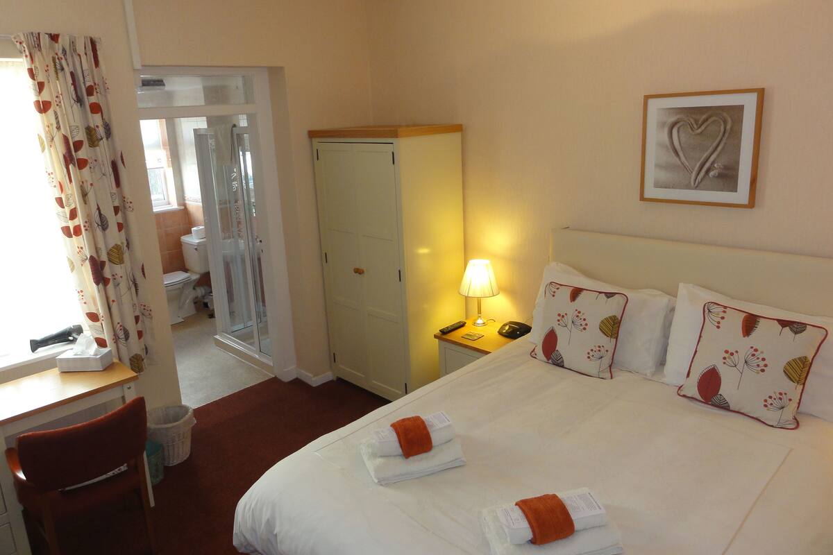 A hotel room with a bed, a desk, a chair, a nightstand with a lamp, and a door leading to a bathroom.