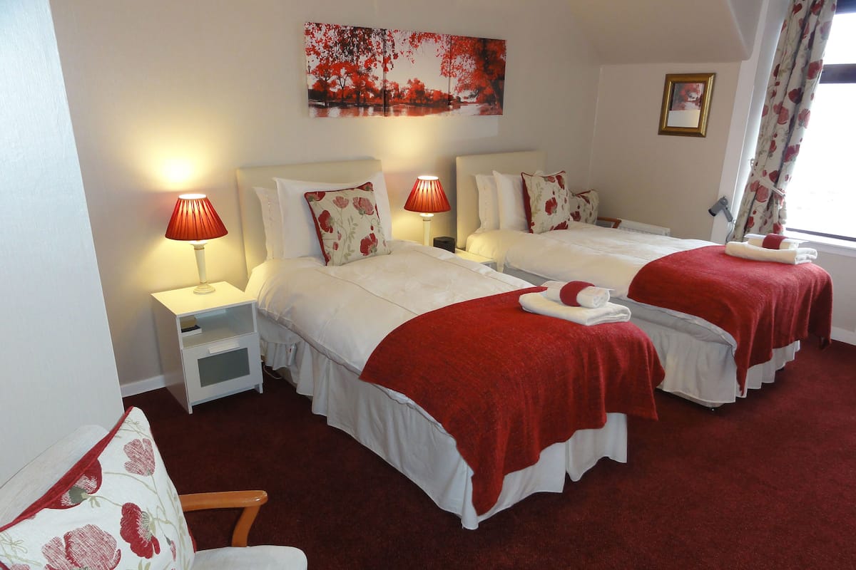 A hotel room with two beds, each with a red blanket and pillows, white bedding, and a red lamp on a bedside table.