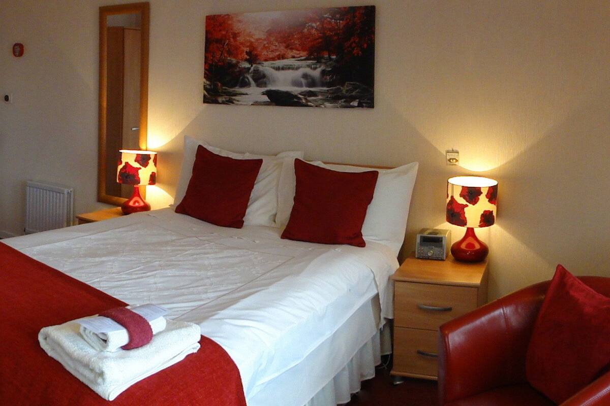 A hotel room with a bed, a nightstand with a lamp, a red chair, and a painting on the wall.