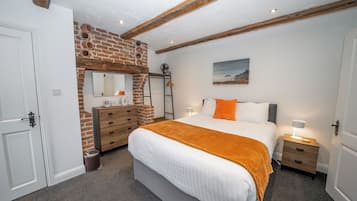 Ein Schlafzimmer mit Backsteinwand, einem Bett mit weißer Bettwäsche und orangefarbenen Kissen, einem Schminktisch, einem Nachttisch mit Lampe und einem Bild an der Wand.