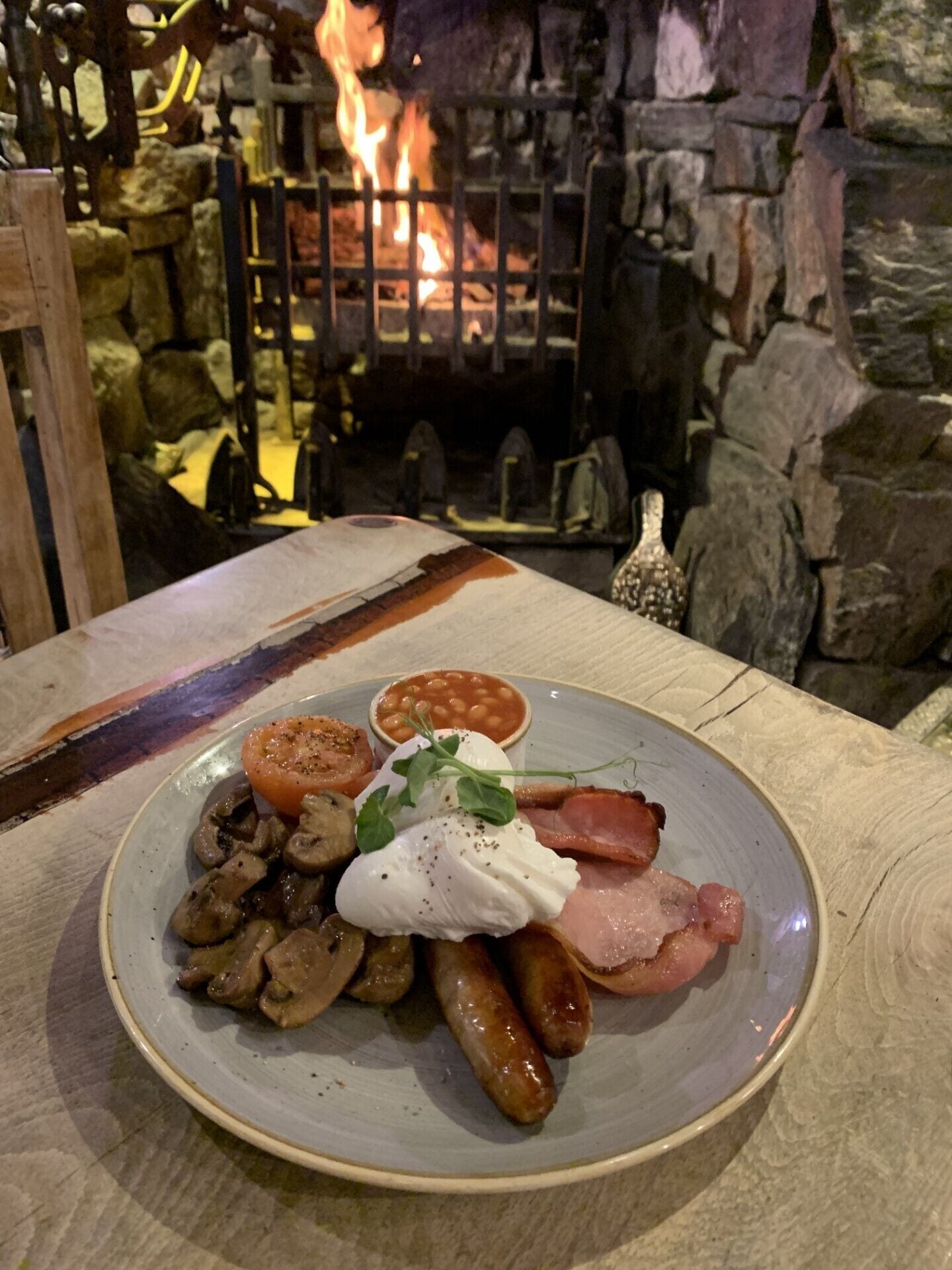Un plato de comida para el desayuno con huevos, tocino, salchichas, champiñones y frijoles, con una chimenea de piedra al fondo.
