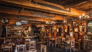 A rustic interior with wooden tables and chairs, a bar area with shelves of bottles, and a stone wall. - The Oak Tree Inn (Balmaha)