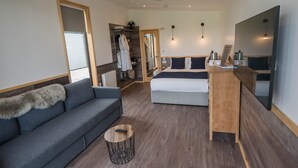 A modern hotel room with a grey sofa, a wooden bedside table, a bed with a dark headboard, a large mirror, and a TV. - The Oak Tree Inn (Balmaha)