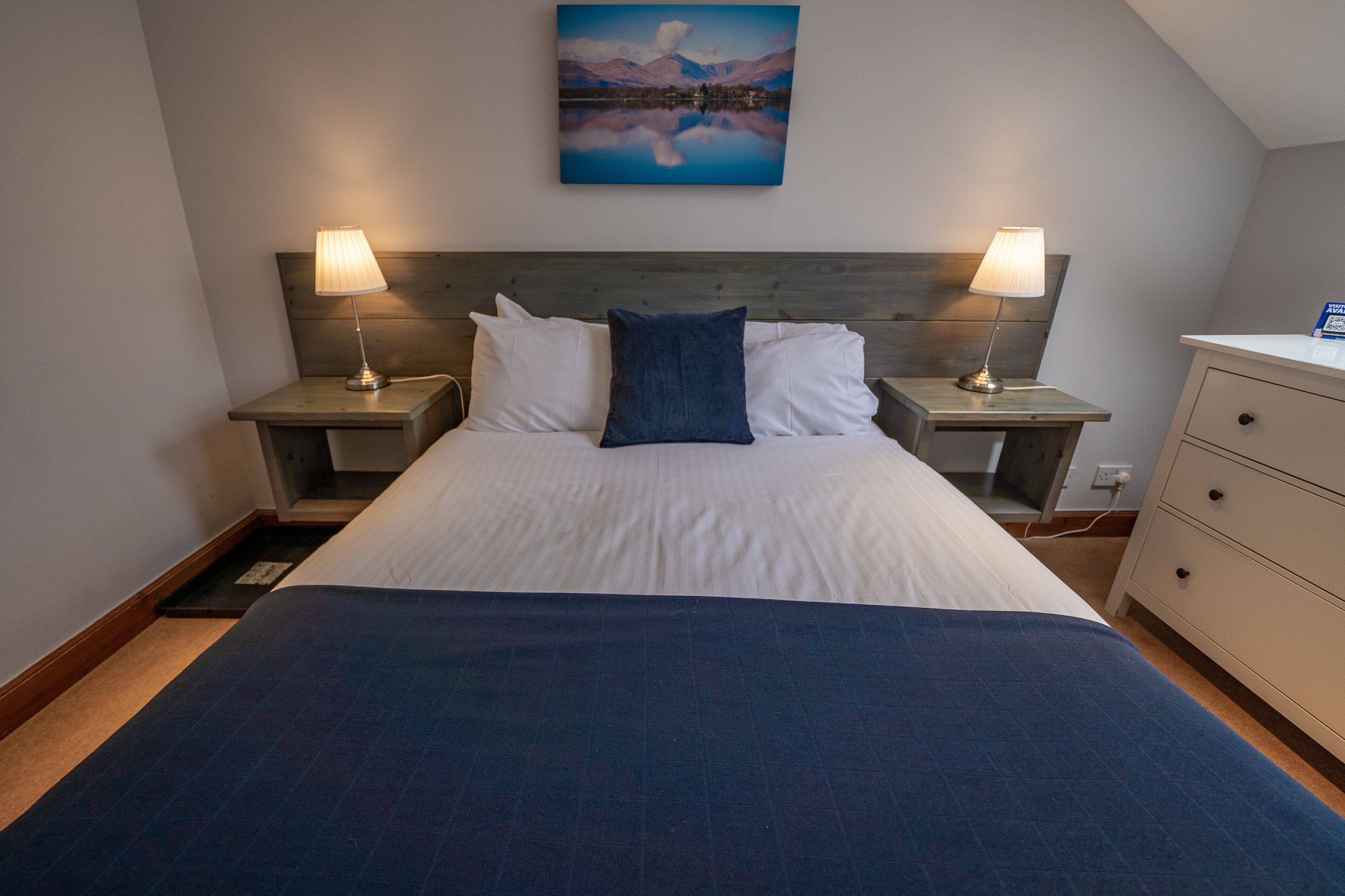 Un lit bien fait, avec une couverture bleue, deux lampes de chevet, une tête de lit et un tableau au-dessus du lit.