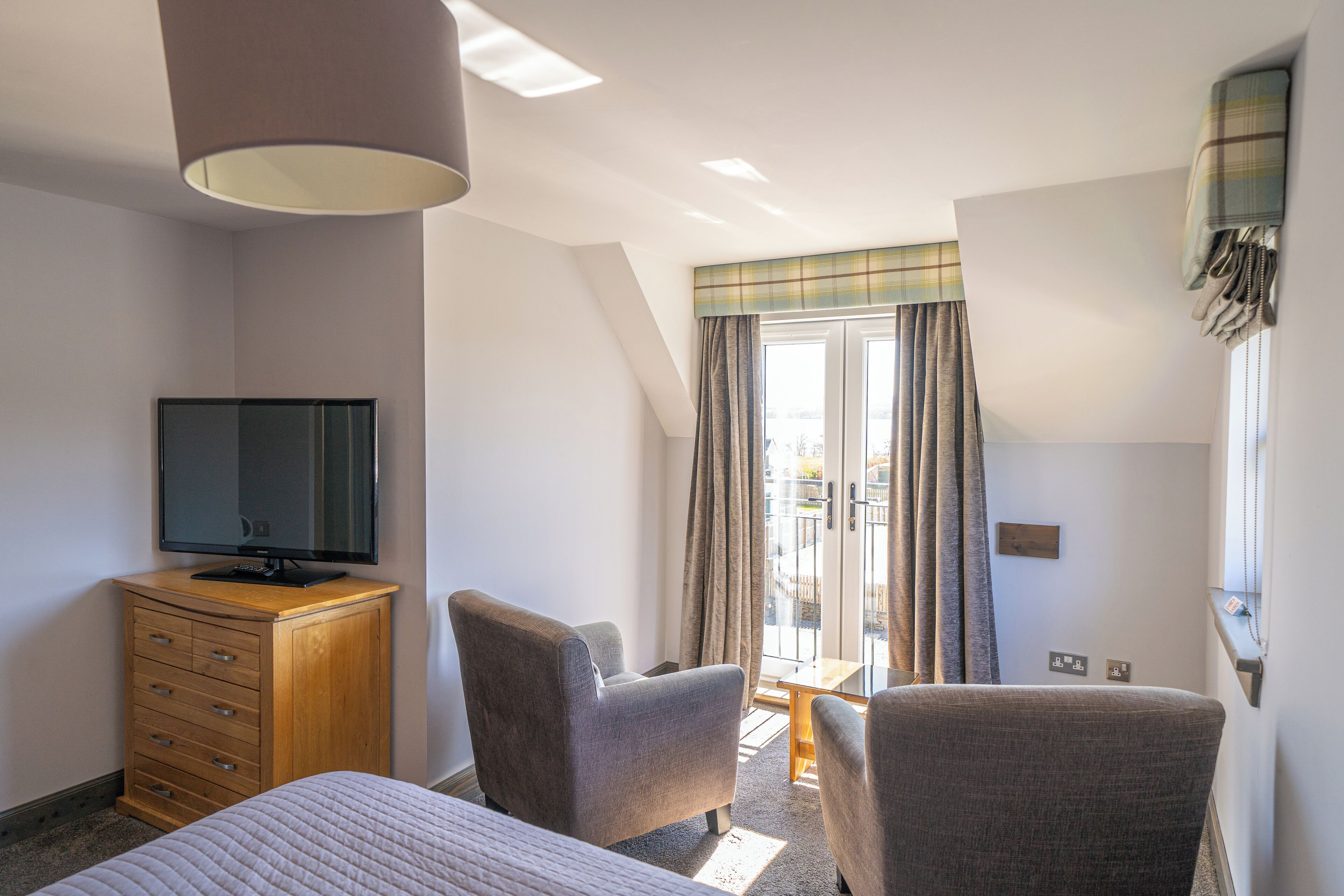 Habitación de hotel con cama, televisor sobre una cómoda de madera, dos sillones, una mesita y una ventana con cortinas.