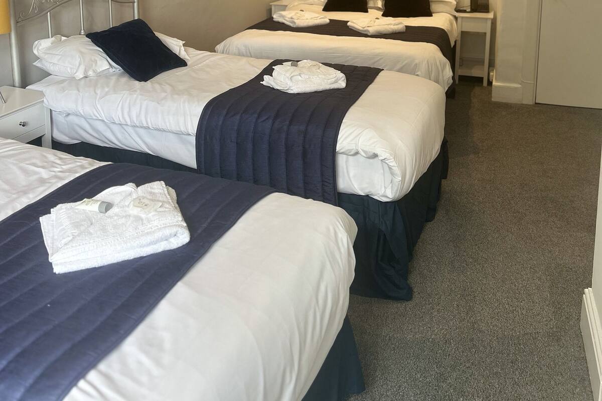 A hotel room with three single beds, each with white bedding and dark headboards. There are bedside tables with lamps and a small nightstand with a phone. The room has a carpeted floor and a door with a sign.