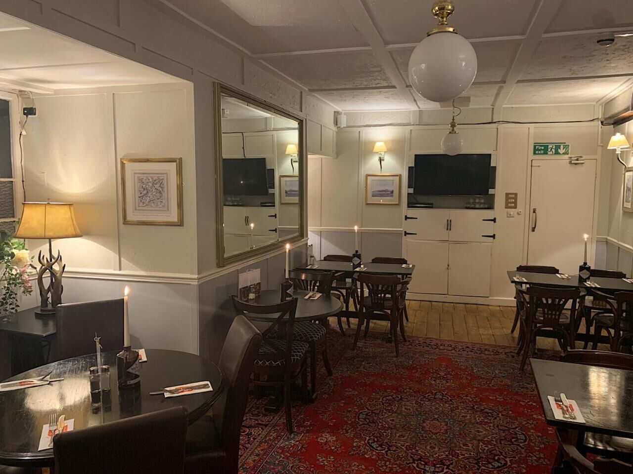 A hotel dining area with tables, chairs, a red carpet, a television, and a fire exit sign.