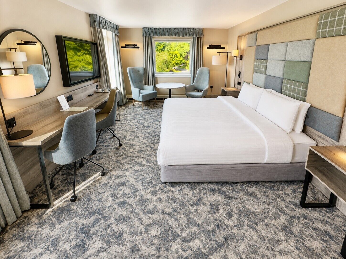 Quarto de hotel com uma cama grande, uma escrivaninha, duas cadeiras, uma televisão e uma janela com vista para árvores.