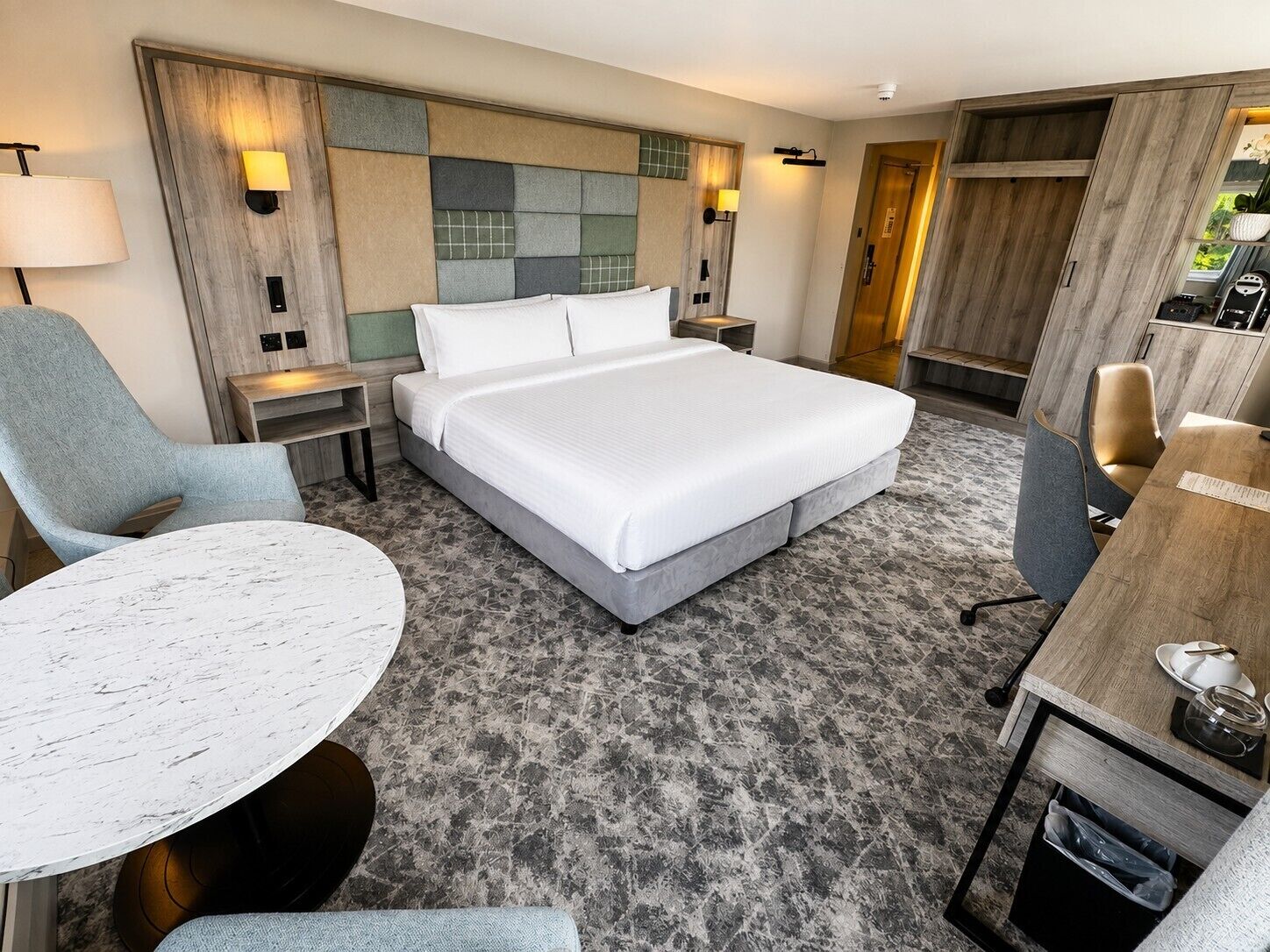 A hotel room with a large bed, a round table, a chair, a desk, and a wardrobe.