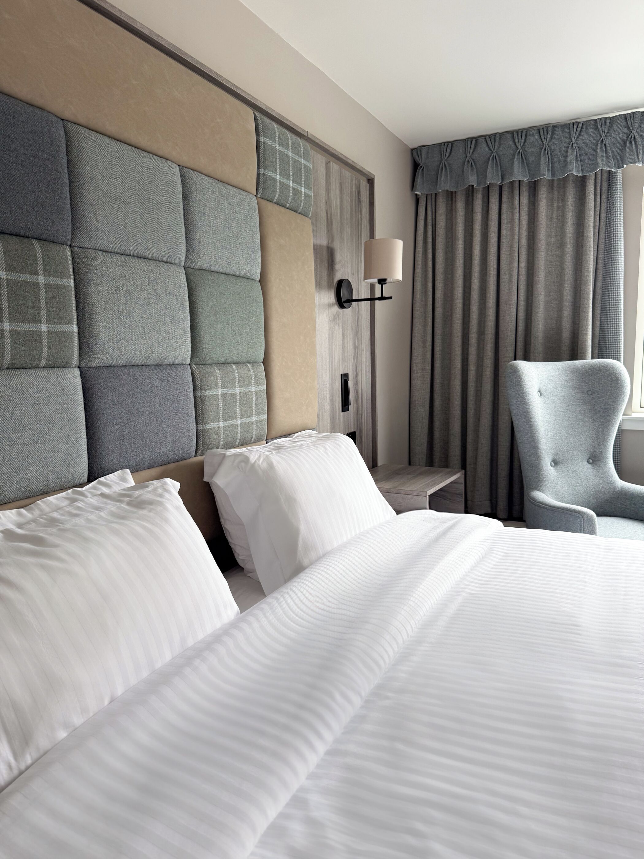 A hotel room with a large bed, a grey armchair, a wall-mounted lamp, and a window with curtains.