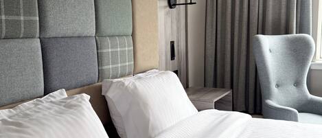 A hotel room with a large bed, a grey armchair, a wall-mounted lamp, and a window with curtains.