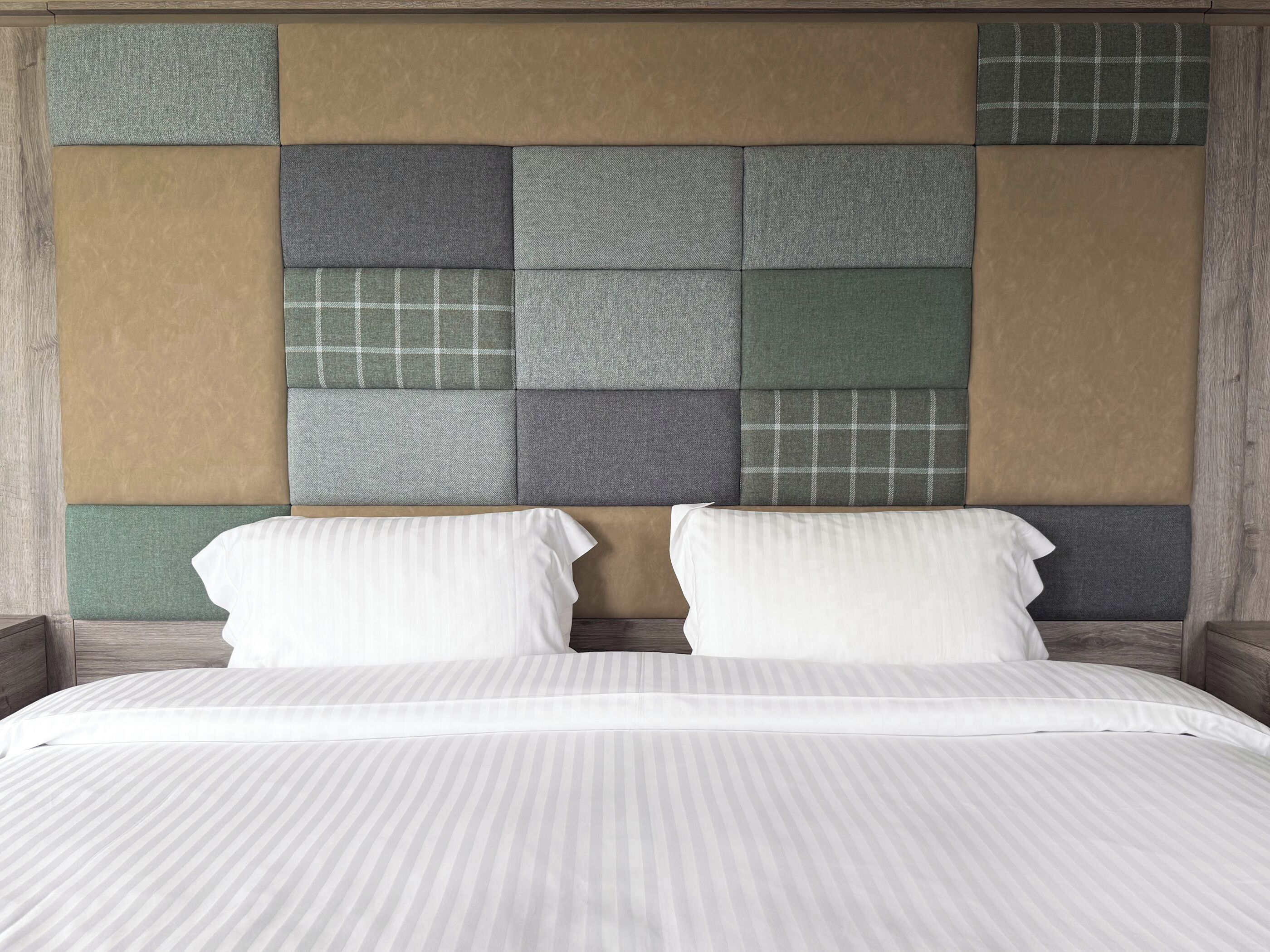 A bed with a white striped bedspread and two white pillows, set against a headboard with a variety of textured panels in neutral tones.