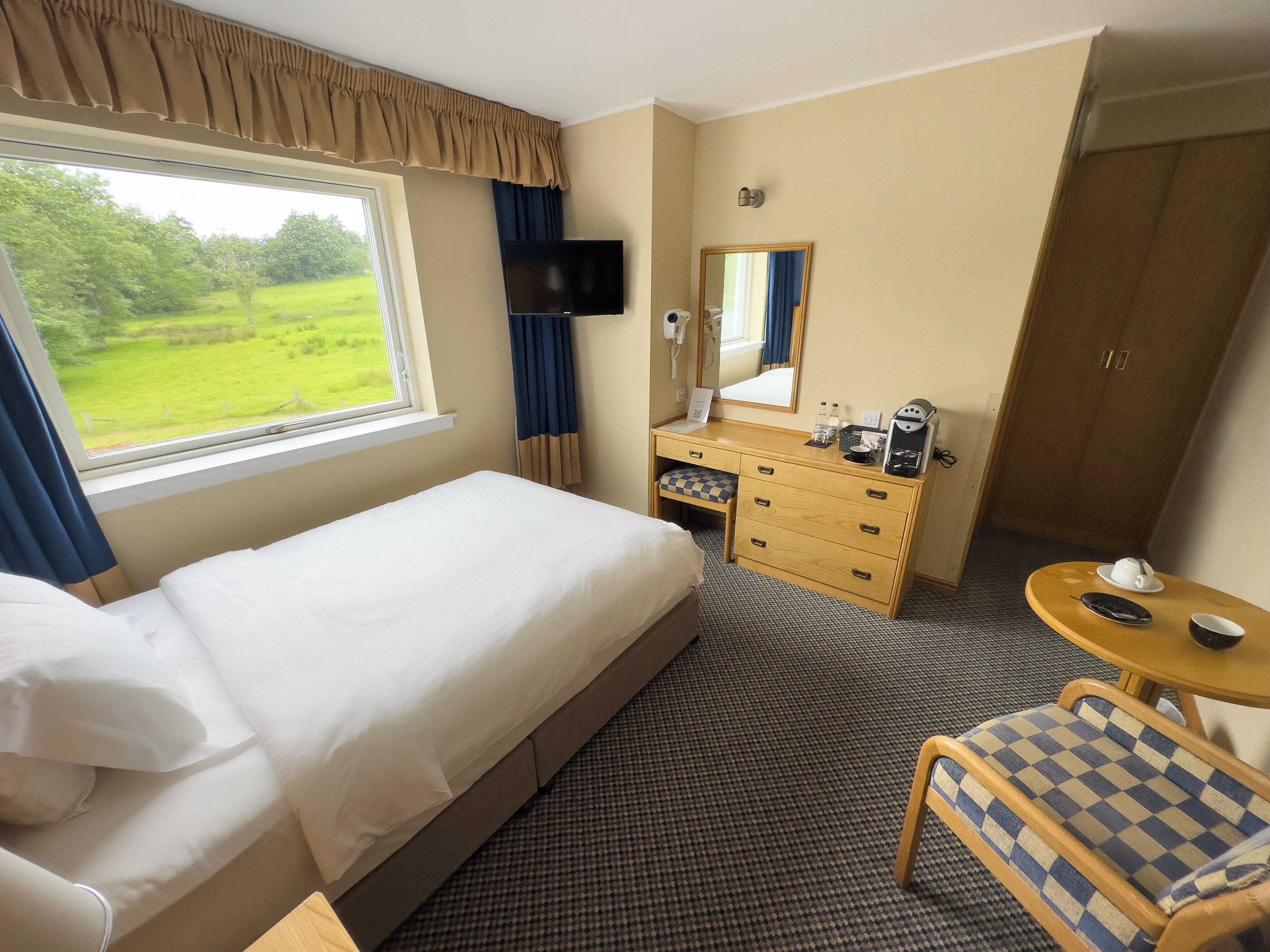 A hotel room with a bed, a desk with a mirror, a chair, and a view of a green landscape.