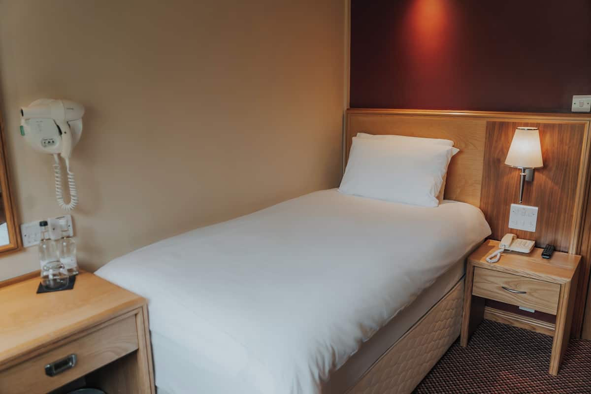 A hotel room with a single bed, a bedside table with a lamp, a phone, and a hairdryer on the wall.