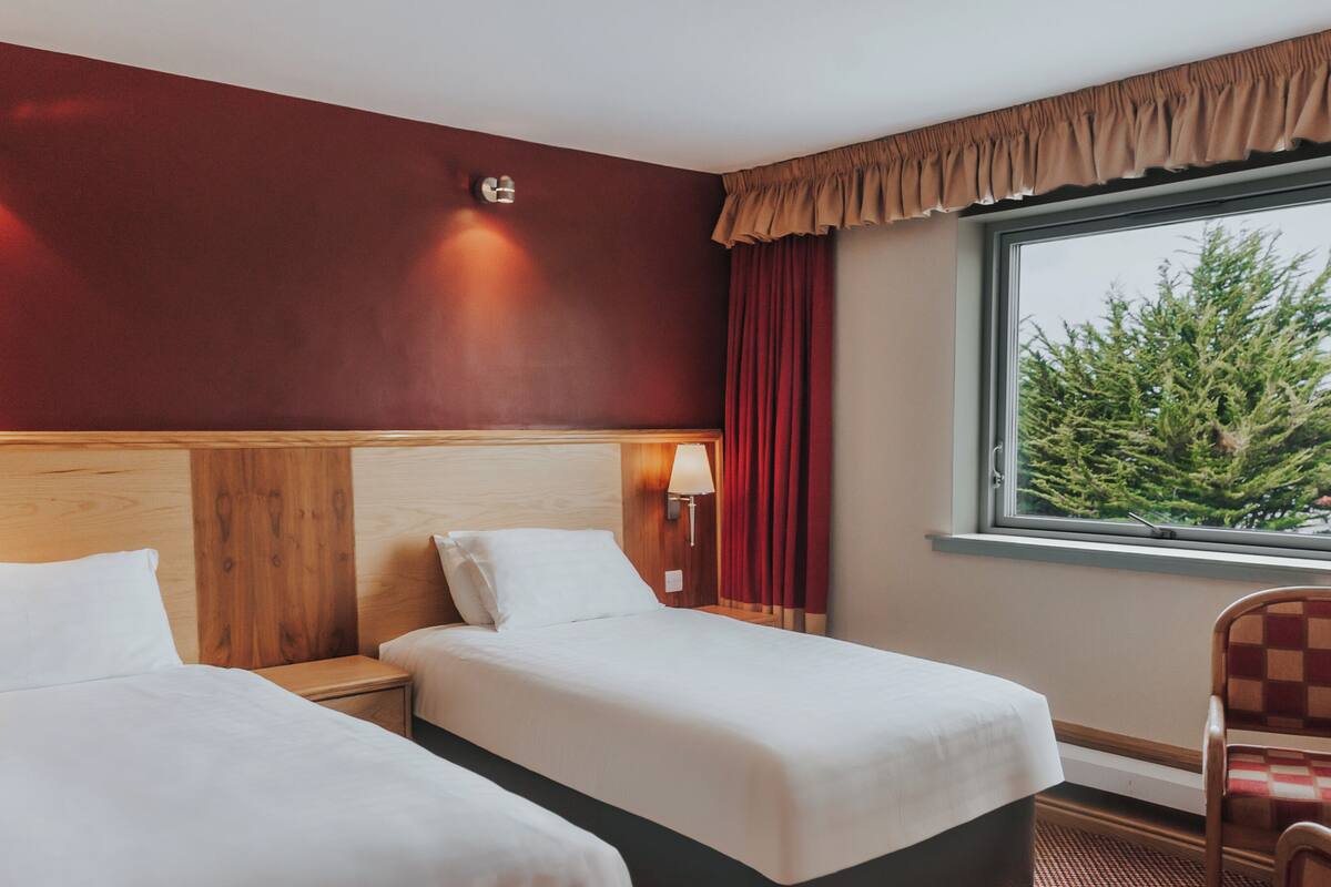 A hotel room with two beds, a wooden headboard, a window with a view of trees, and a checkered chair.