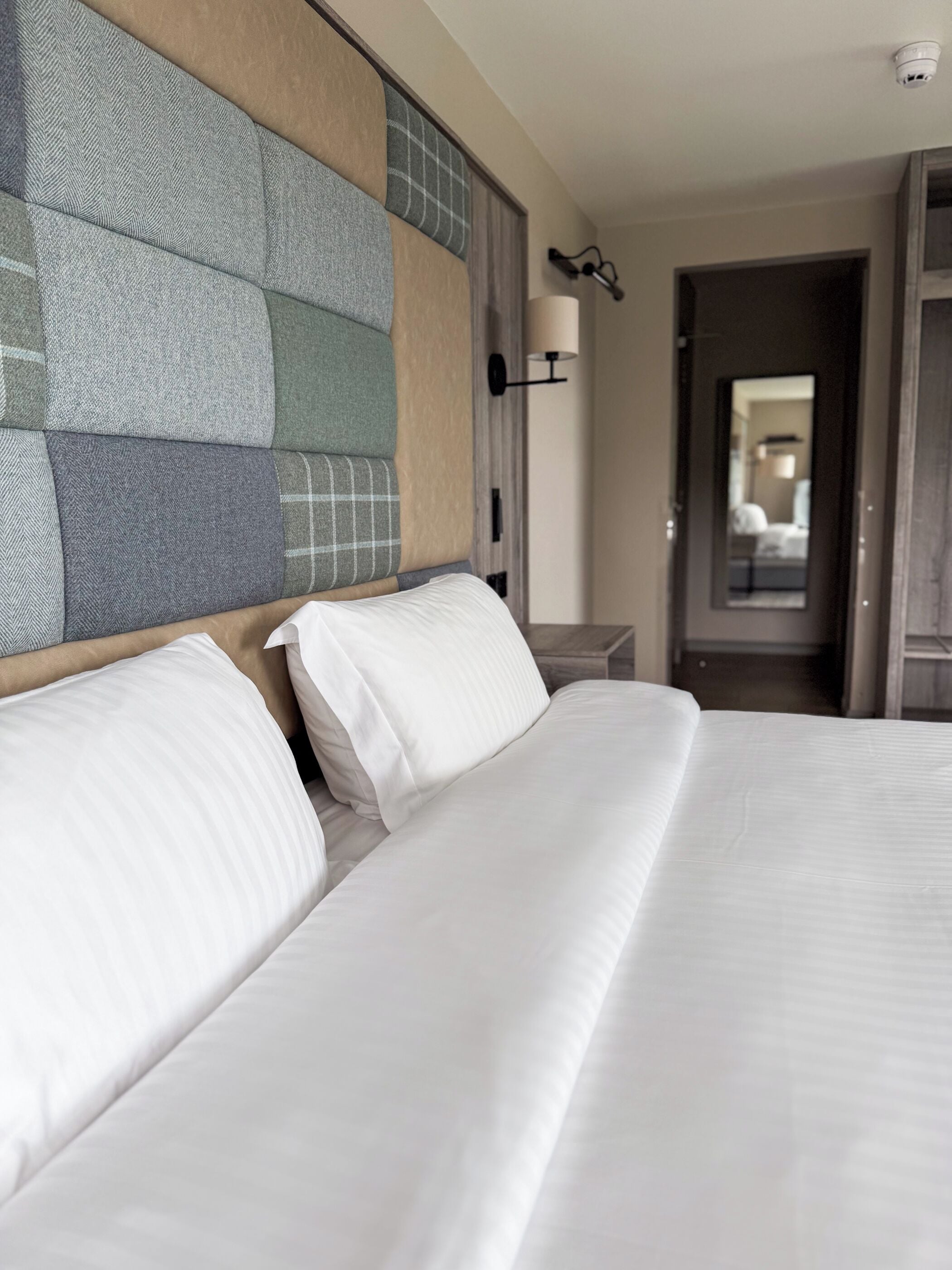 Quarto de hotel com uma cama grande, cabeceira com padrão geométrico, mesa de cabeceira com abajur e espelho na parede.