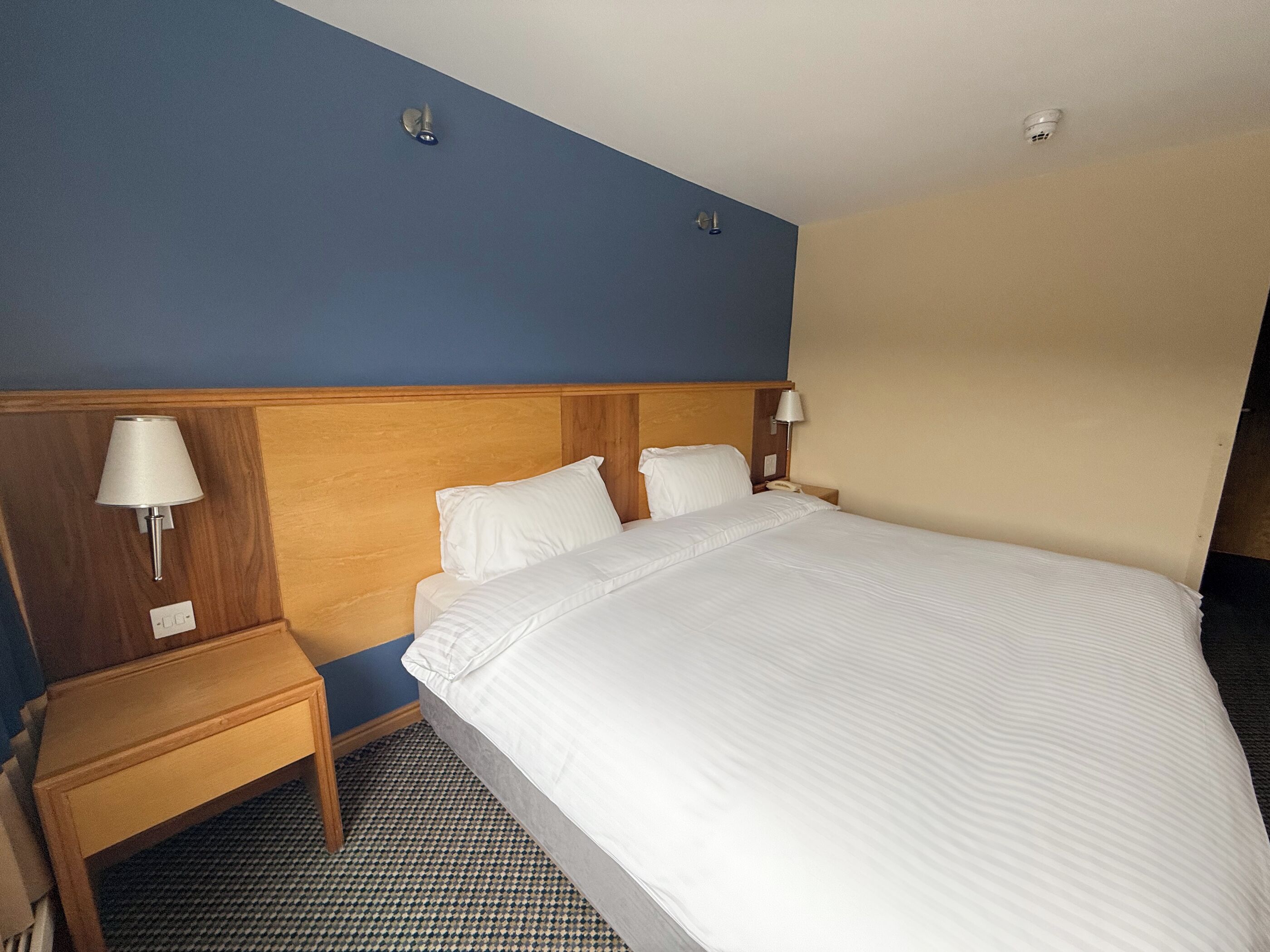 Una habitación de hotel con una cama grande, dos mesitas de noche y dos lámparas fijadas a la pared.