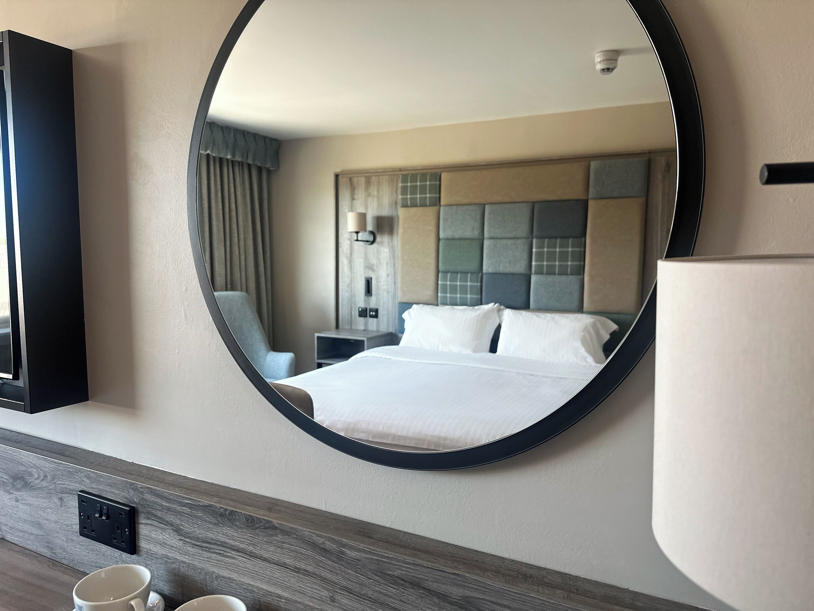 Une chambre d’hôtel avec un grand miroir rond, un lit recouvert de draps blancs, une table de chevet avec une lampe, une chaise et une petite table en bois sur laquelle sont posées deux tasses.