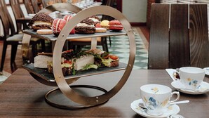 A three-tiered dessert stand with various pastries and sandwiches, a white floral cup and saucer, and a wooden table. - The Cabarfeidh Hotel (Stornoway)