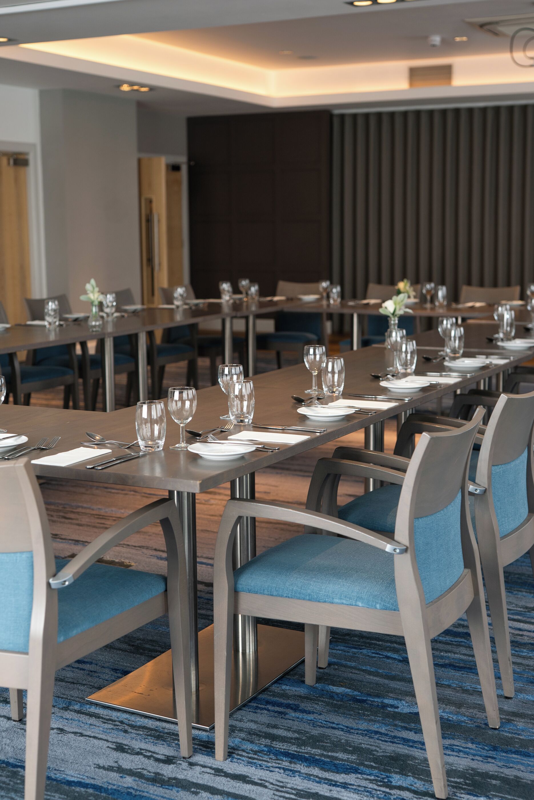 Une salle à manger d’hôtel avec de longues tables dressées pour le service, des chaises dotées de coussins bleus et un sol recouvert de moquette.