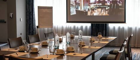 Een vergaderruimte met een lange tafel gedekt voor een vergadering, een scherm waarop tekst wordt geprojecteerd en een groot raam met gordijnen.