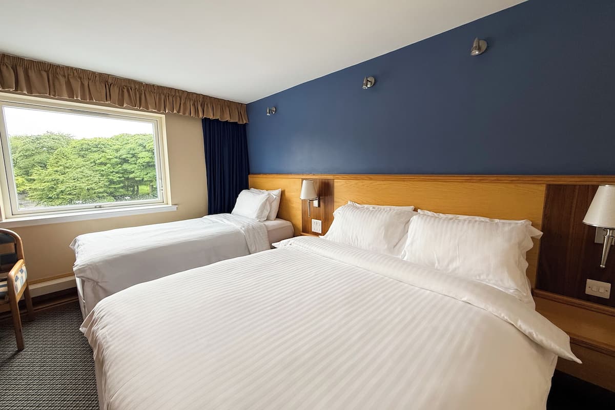 A hotel room with two beds, a window with a view of greenery, a wooden headboard, and a nightstand with a lamp.