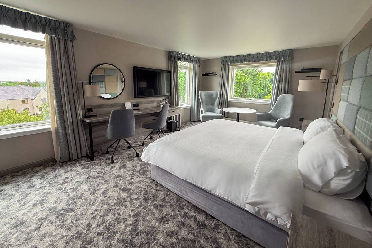 A hotel room with a large bed, two armchairs, a desk with a TV, and a window with a view of buildings and trees.
