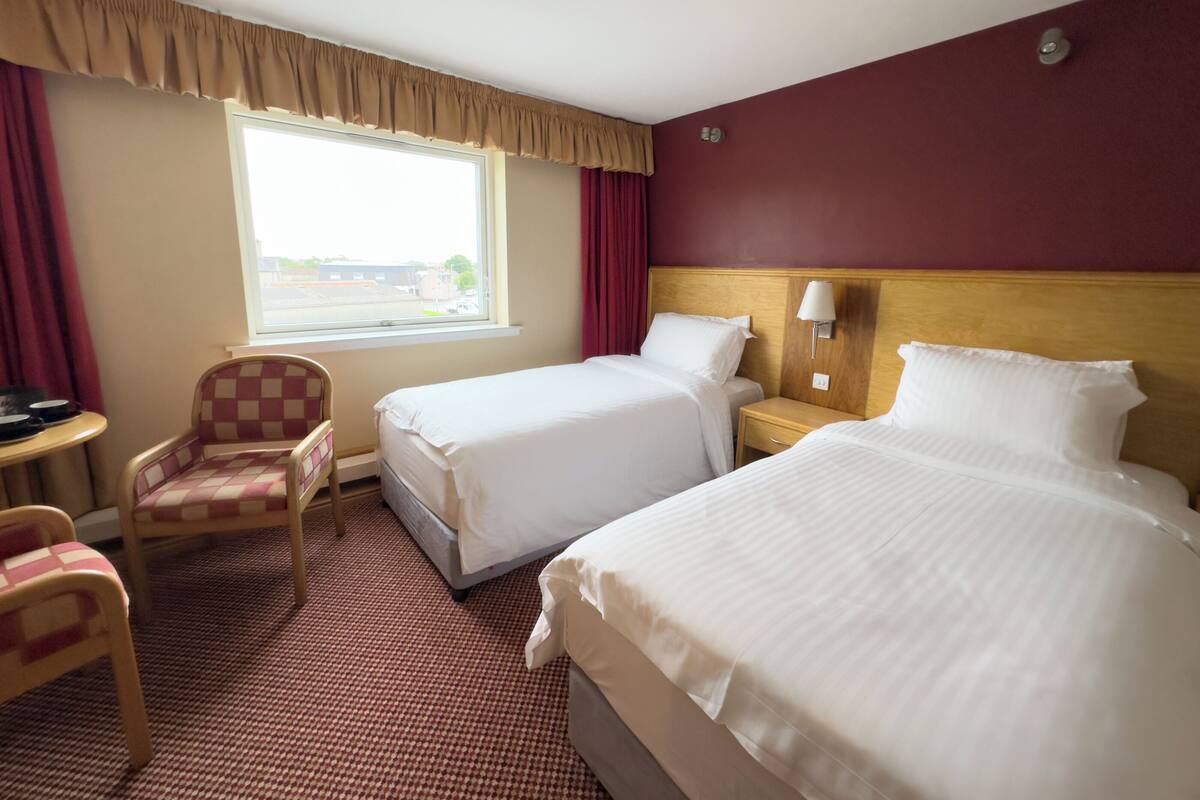 A hotel room with two beds, a chair, a nightstand, and a window with curtains.