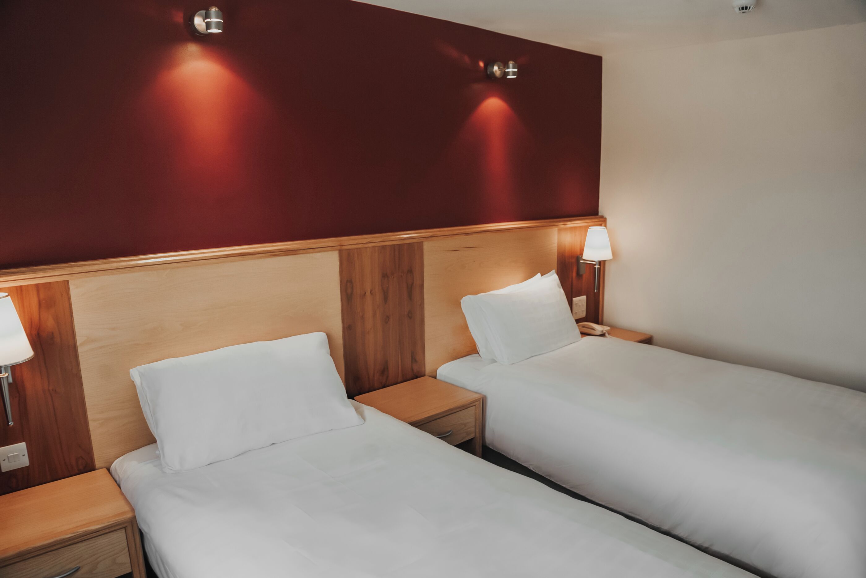 Habitación de hotel con dos camas individuales, cabeceros de madera, lámparas de noche y una pared de acento roja.