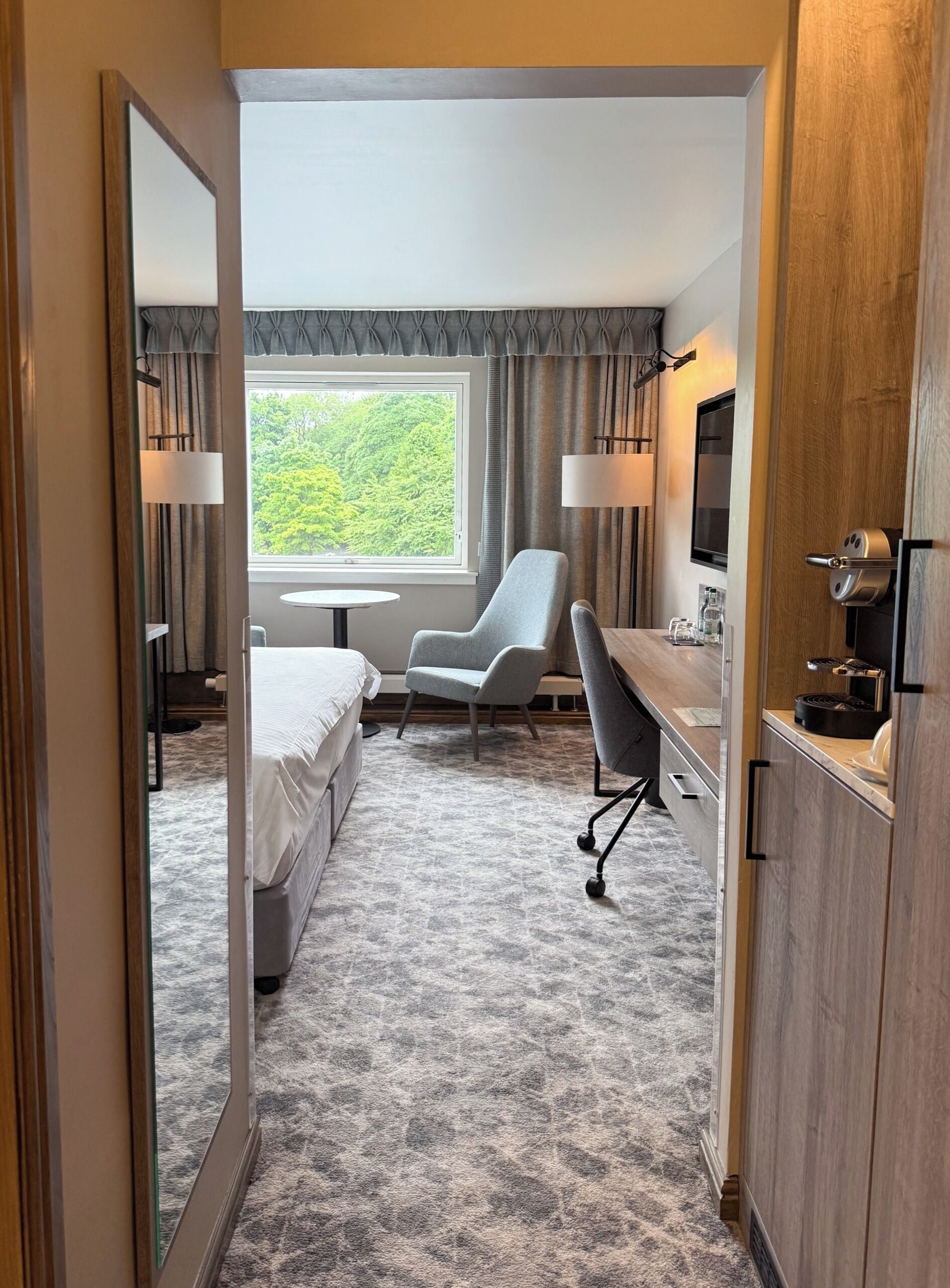 Une chambre d’hôtel équipée d’un lit, d’un bureau, d’une chaise, d’une télévision et d’une fenêtre donnant sur la verdure.