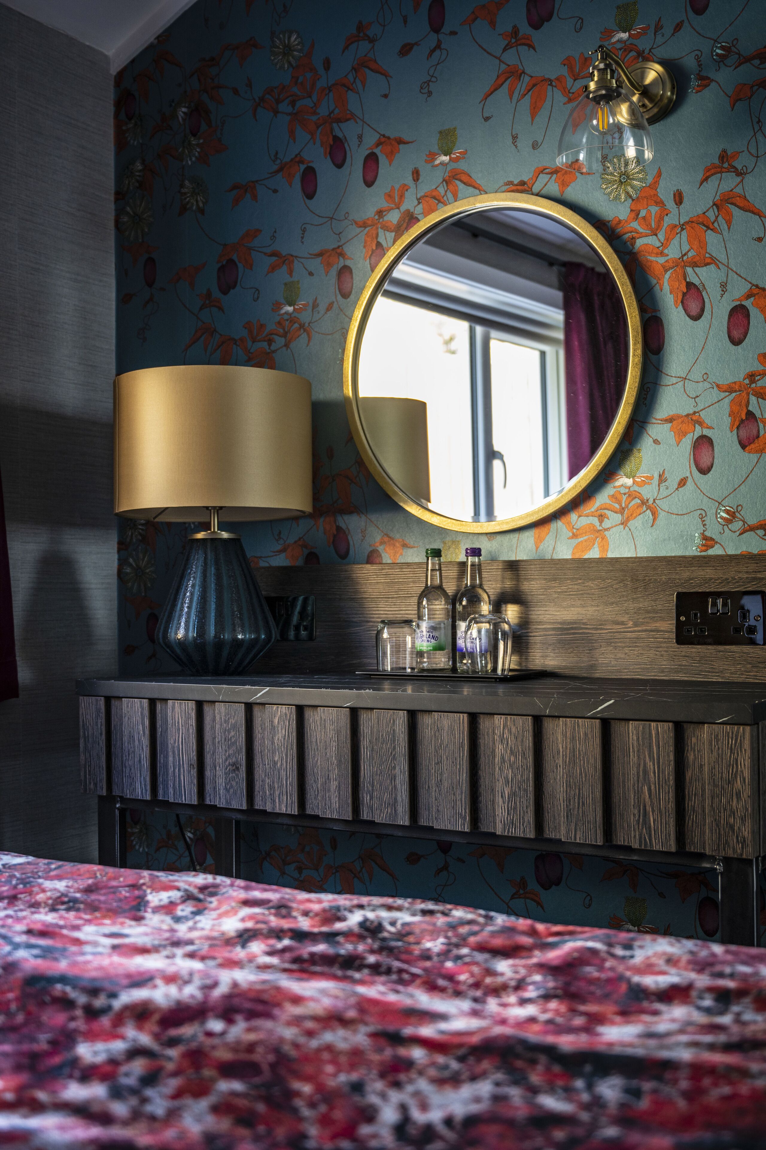 A bedroom with a wooden dresser, a round mirror, a lamp, and a floral wallpaper.