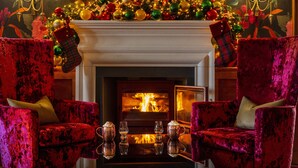 A fireplace with a Christmas tree and stockings, flanked by two red velvet armchairs with a tray of drinks in front.