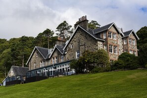 Situated on a grassy slope, this traditional building features stone walls and a pitched roof, surrounded by lush greenery and trees.