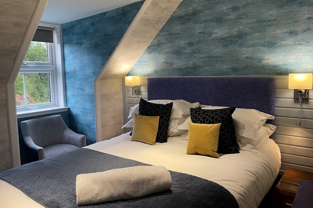 Une chambre d’hôtel moderne avec un plafond en pente, un grand lit avec plusieurs oreillers, une chaise et deux lampes de chevet.