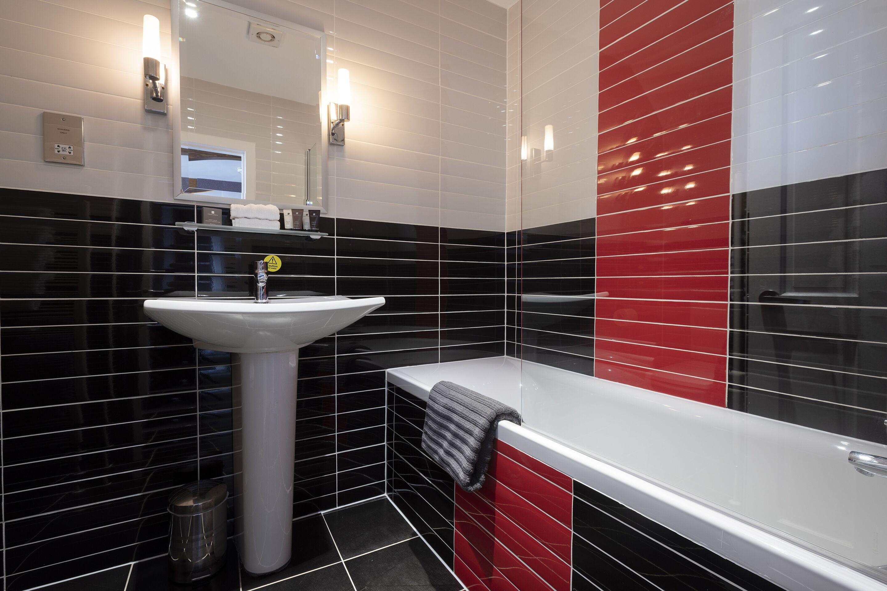 A modern bathroom with a white sink, a mirror, and a red and black tiled wall.