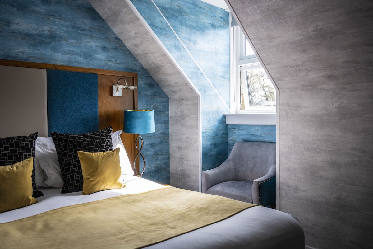 Une chambre d’hôtel moderne avec un plafond en pente, un lit agrémenté d’oreillers à motifs, une lampe bleue et un fauteuil.