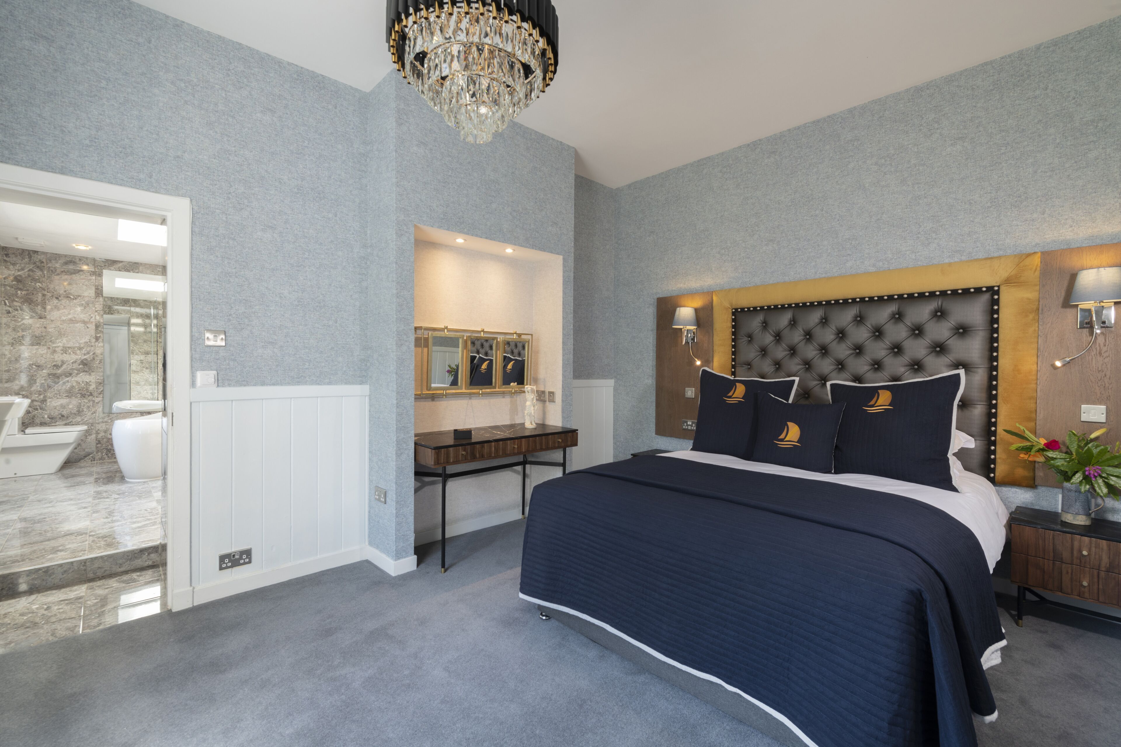 A bedroom with a large bed, a chandelier, a vanity with a mirror, and a bathroom visible through an open door.