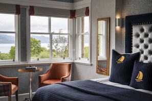 Relax in a serene room with a large bed, tufted headboard, and armchairs, offering a view of a body of water and greenery.
