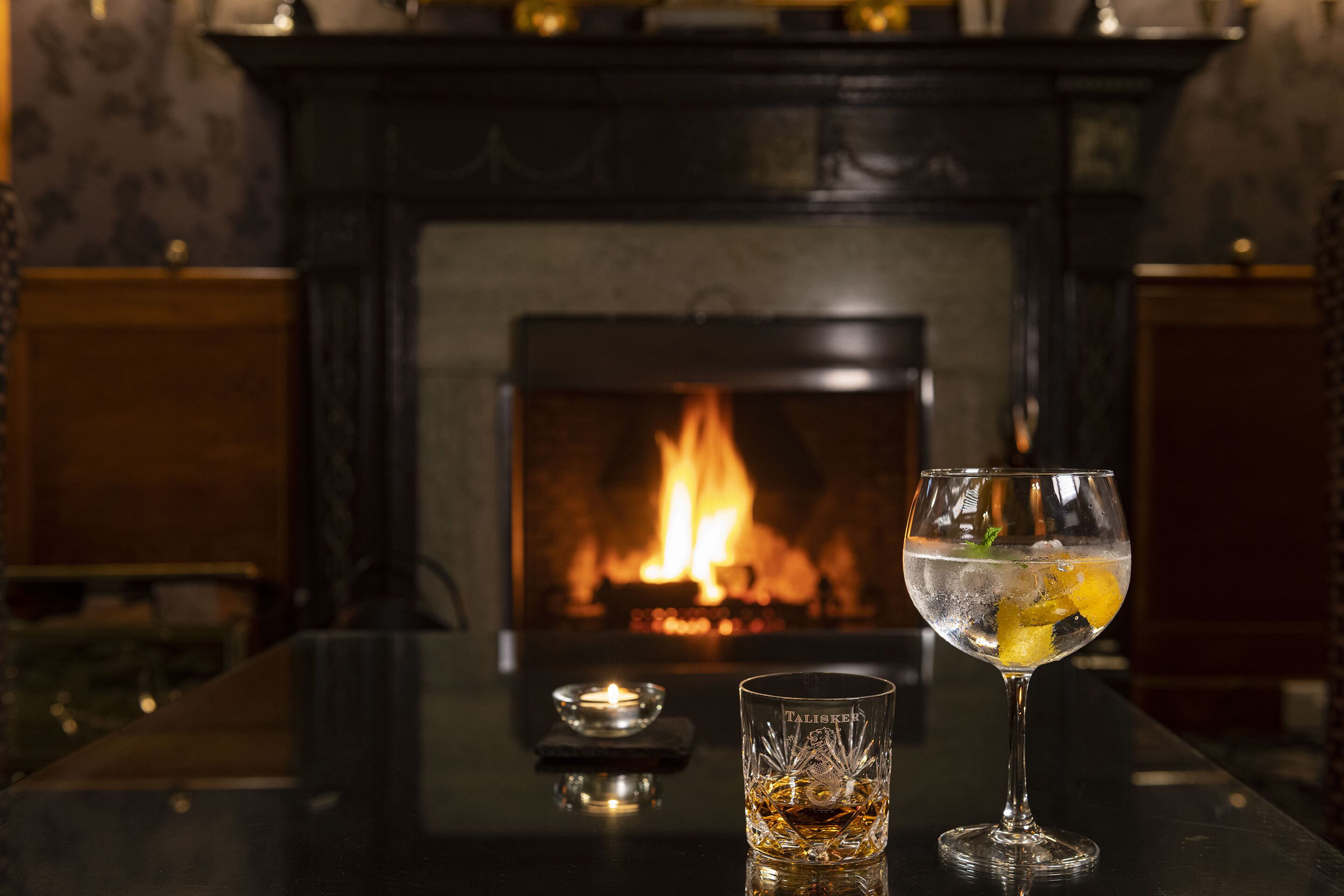 A glass of a clear beverage with a garnish, a candle, and a fireplace in the background.