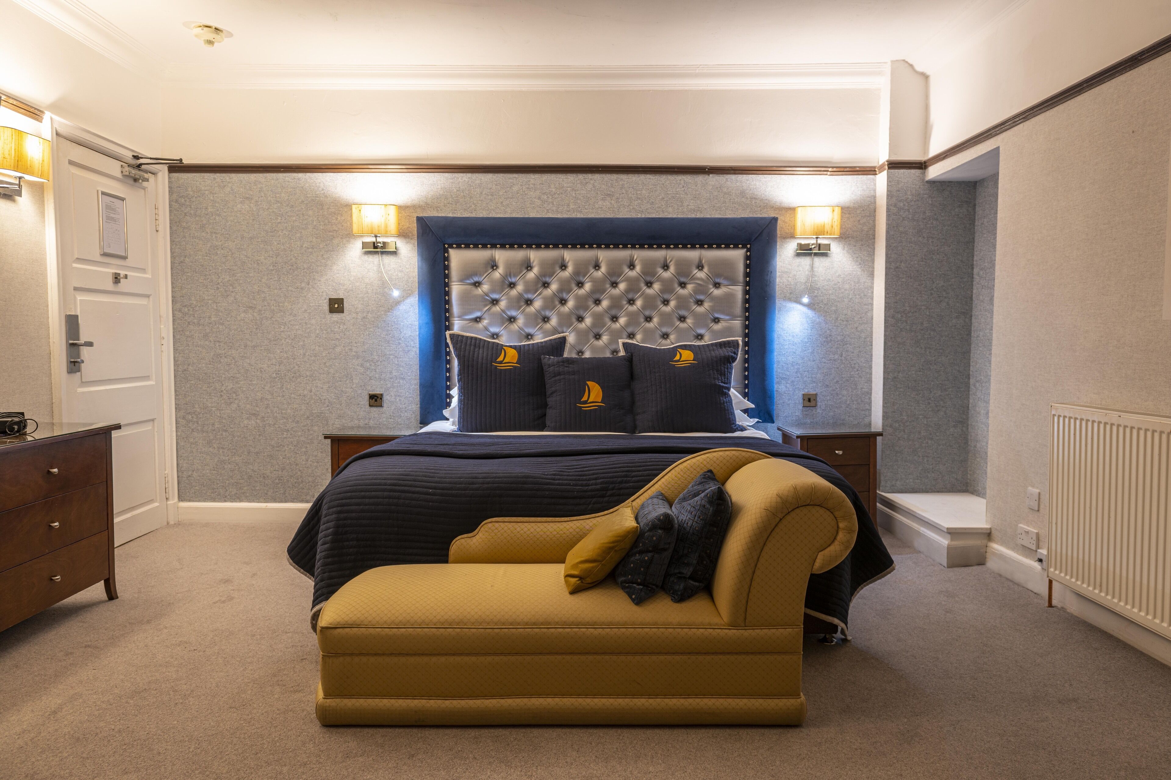 Quarto com cabeceira estofada, cama com roupa de cama escura, poltrona amarela e abajures.