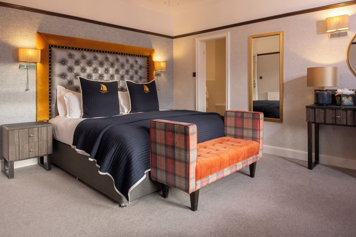 Une chambre d’hôtel avec un grand lit, un fauteuil à motifs écossais, une table de chevet et une commode.
