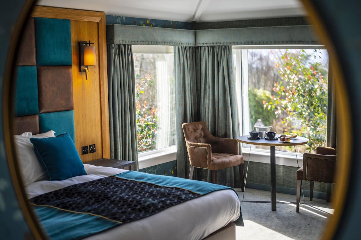 Une chambre d’hôtel avec un lit, une chaise, une table avec une lampe et une fenêtre avec des rideaux.