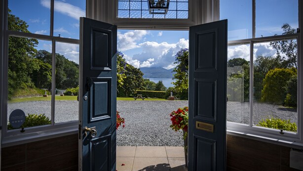 A view through a large open door with multiple windows, leading to a gravel area with a bench and a statue, surrounded by greenery and a mountain in the distance.