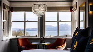 A hotel room with a large bed, two red armchairs, a small round table, a chandelier, and a view of mountains and a lake through large windows.