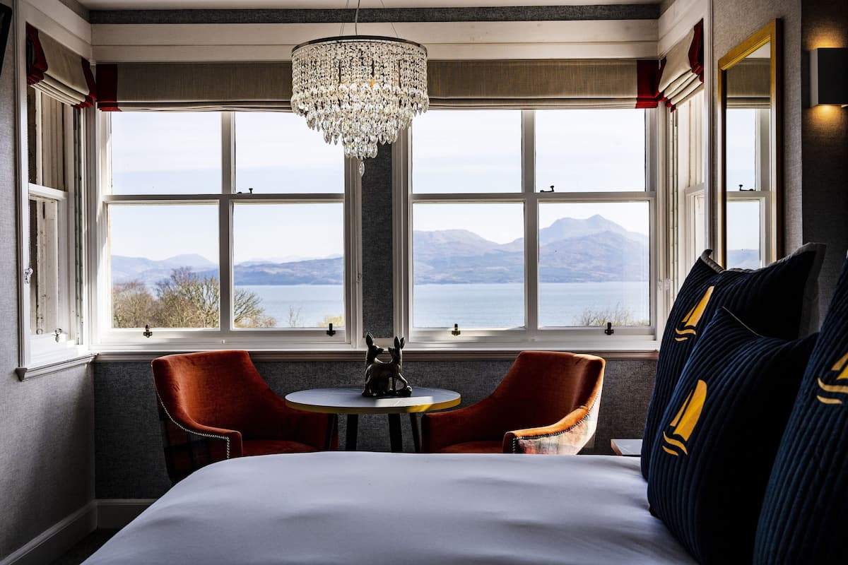 Une chambre d’hôtel avec un grand lit, deux fauteuils rouges, une petite table ronde, un lustre et une vue sur les montagnes et un lac, visible à travers de grandes fenêtres.